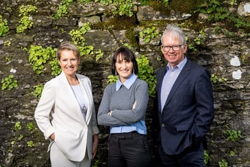 Two white women and one white male standing against a wall, smiling.