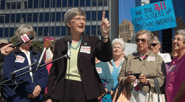 Woman in black suit with white short hair speaking passionately to other women.